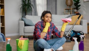 woman in rubber gloves sits on floor with cleaning supplies perplexed expression