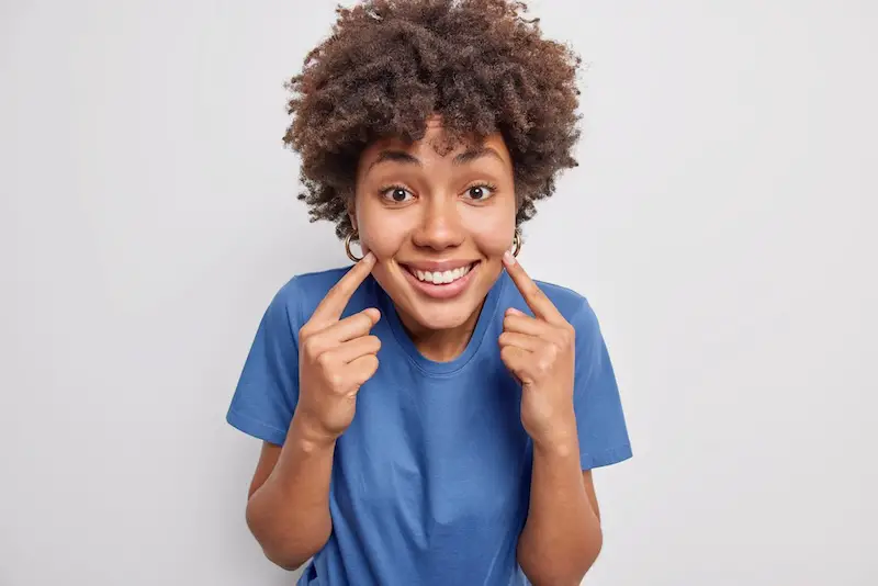 young woman points index fingers at cheeks smiles dental health