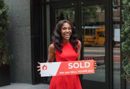 Smiling woman holding just sold sign outside real estate office