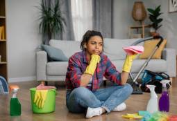 woman in rubber gloves sits on floor with cleaning supplies perplexed expression