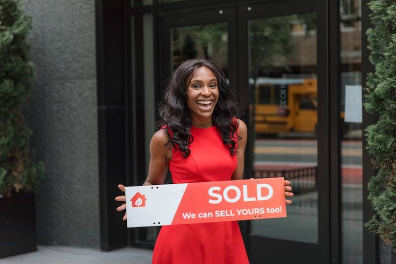 Smiling woman holding just sold sign outside real estate office