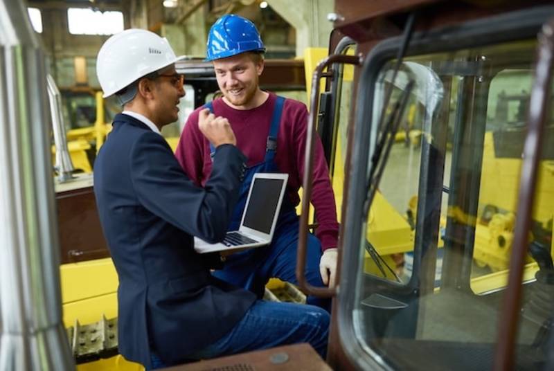 man-in-suit-helmet-laptop-talking-to-forklift-driver.