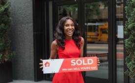 Smiling woman holding just sold sign outside real estate office