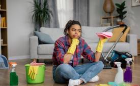woman in rubber gloves sits on floor with cleaning supplies perplexed expression